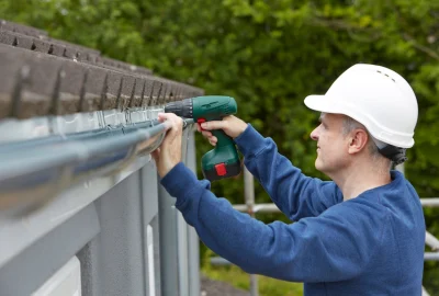 A construction worker in a white hard hat uses a green cordless drill to install a gutter on a house.