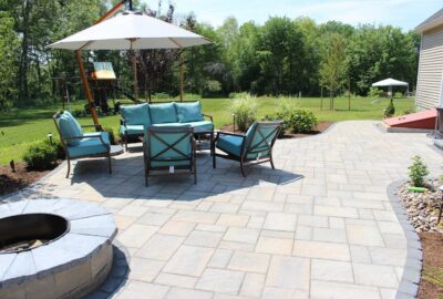 Patio with light gray pavers, teal outdoor furniture under a white umbrella, and a stone fire pit in a grassy yard.