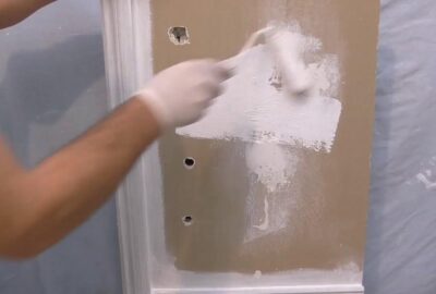 A person in white gloves uses a paint roller to apply white paint to a tan door.