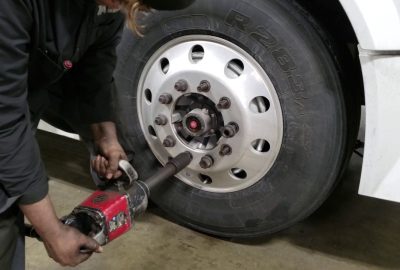 A mechanic uses a red pneumatic impact wrench to tighten lug nuts on a large truck wheel.