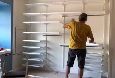 Man in a yellow t-shirt installing white shelving and hanging rods in a walk-in closet.