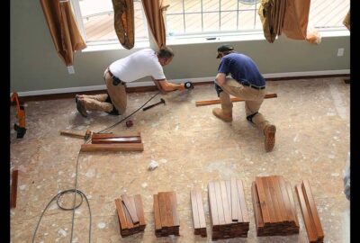Two men installing hardwood flooring on a subfloor in a room with large windows.