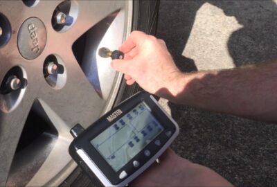 Hand using a Masten TPMS tool to program a tire pressure sensor on a Jeep wheel.
