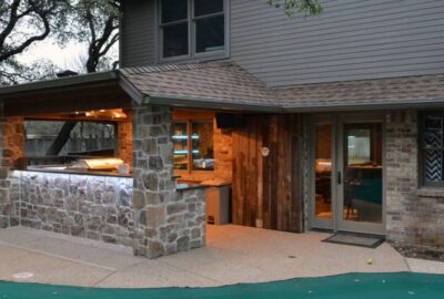 Covered outdoor kitchen and bar with a stone countertop and integrated lighting attached to a house.