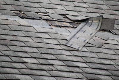 Damaged gray asphalt roof shingles with torn and lifted sections exposing the wooden underlayment.
