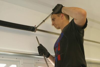 Man in black gloves using metal rods to adjust a garage door torsion spring.