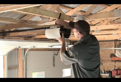 Man installing a white garage door opener motor to wooden ceiling beams in a garage.