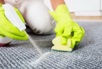 Hands in yellow gloves spraying cleaner and scrubbing a gray textured carpet with a sponge.