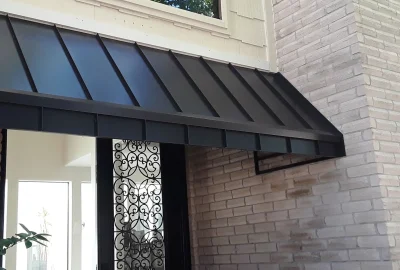 Black metal awning installed above a front door with ornate iron grillwork and light-colored brick siding.