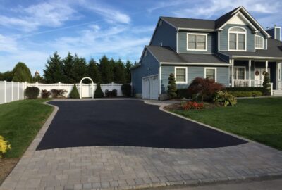A wide asphalt driveway with a gray stone paver entrance leads toward a blue two-story house with a white fence.