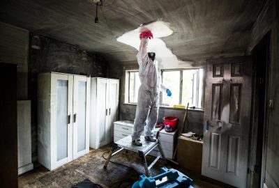 Worker in white protective gear on a ladder repairing a ceiling in a room under renovation.