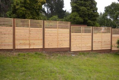 Modern wooden fence with horizontal light-wood panels and a slatted upper section, backed by green trees.