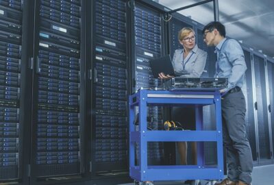 Two IT professionals discuss data on a laptop next to a server rack and a blue equipment cart in a data center.
