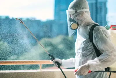 Person in protective hazmat suit spraying disinfectant using a backpack sprayer on a balcony.