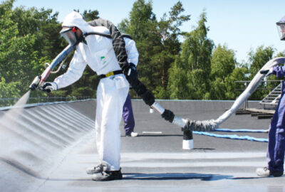 Two workers in protective hazmat suits spray a coating onto a flat rooftop with specialized equipment.