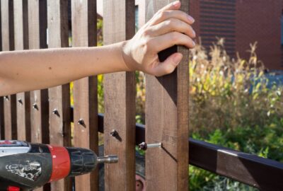 A person uses a red and black power drill to screw a bolt into a vertical wooden fence slat.