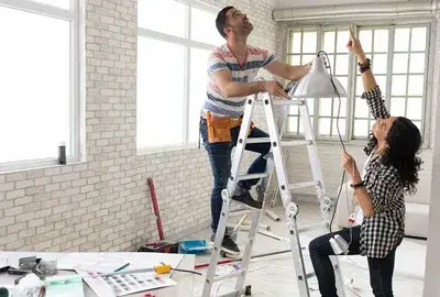 A man on a ladder and a woman work together to install a white pendant light in a bright, modern room.