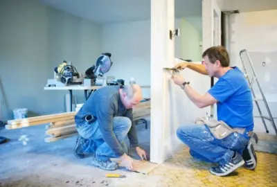 Two carpenters installing trim in a room under renovation.
