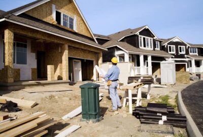 A construction worker in a yellow hard hat reviews blueprints at a residential site with houses under construction.