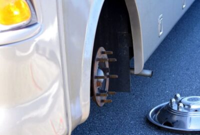 White vehicle missing a wheel, showing the exposed hub and a detached chrome rim on the asphalt.