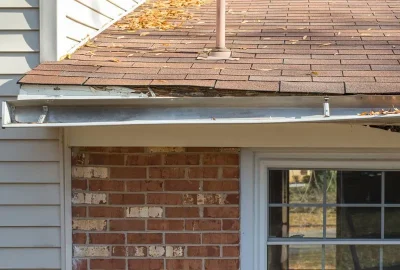 *   Image shows a close-up of a house's roofline.
    *   Key elements: Brown shingled roof with fallen leaves, a white gutte