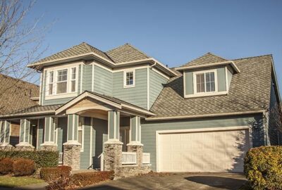 Two-story light blue house with white trim, grey shingled roof, stone porch pillars, and a white garage door.