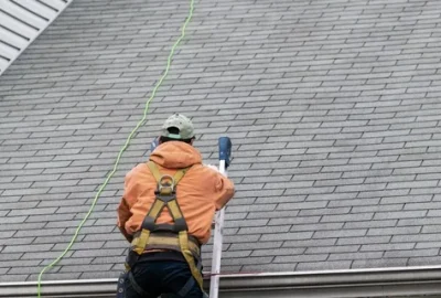 Worker in an orange jacket and safety harness on a ladder working on a grey shingled roof.
