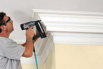 Man using a nail gun to install white crown molding on a ceiling.