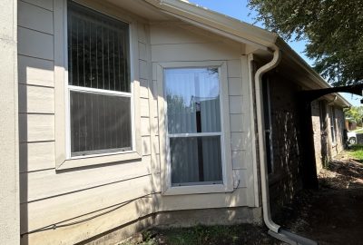 Exterior view of a light-colored house with two windows, white gutters, and a brick wall section.