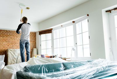 Person painting a white ceiling with a paint roller in a room with furniture covered in plastic sheeting.