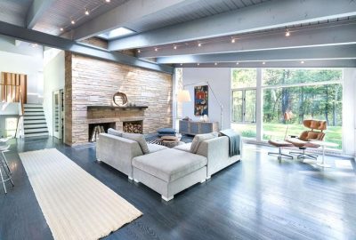 Modern open-concept living room with a grey sectional, stone fireplace, and large windows overlooking a forest.