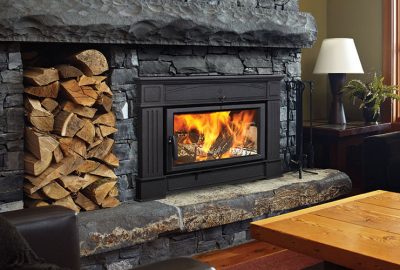 A black wood-burning fireplace with a bright fire inside, built into a grey stone wall with firewood stacked to the left.