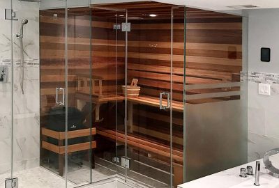 Glass-enclosed sauna with wooden walls and tiered benches, located next to a marble shower.
