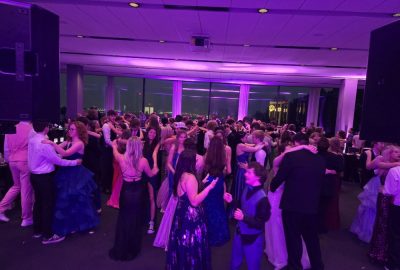 Teens in formal attire dancing in a large ballroom lit with purple light.