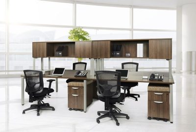 Modern shared office workstation with wooden desks, black mesh chairs, laptops, and overhead storage.
