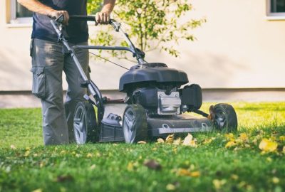 A person pushes a lawn mower across a green yard scattered with yellow autumn leaves.