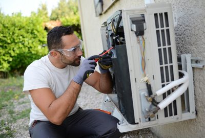 Technician wearing protective goggles and gloves uses a screwdriver to repair an outdoor air conditioning unit.