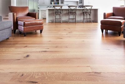 Light-toned hardwood floors in a living room with two leather chairs, matching ottomans, and a kitchen island in the back.
