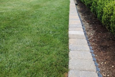 A line of gray stone pavers separating a green lawn from a mulch garden bed with a hedge.