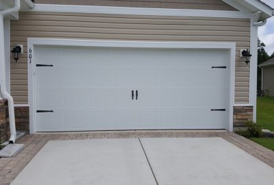White carriage-style double garage door with black decorative hardware on a tan house with a concrete driveway.