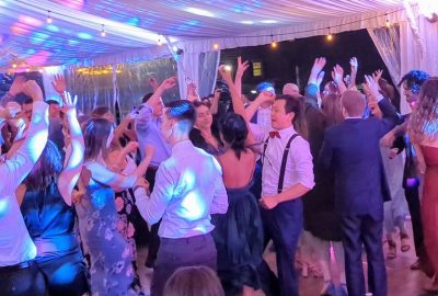 A group of wedding guests dancing on a dance floor under string lights, illuminated by colorful, flashing event lighting.