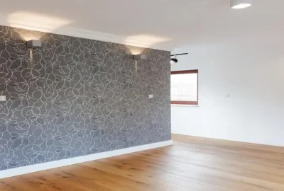 Modern empty room with a grey floral patterned accent wall, light wood floors, and a window in the background.