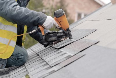 A worker in a safety vest uses a pneumatic nail gun to install grey shingles on a sloped roof.