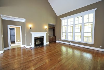 Empty living room with hardwood floors, tan walls, white fireplace, and large windows.