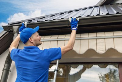 Worker in a blue uniform repairing gutters on a house from a ladder.