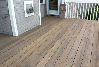 Weathered wooden deck adjacent to a grey-sided house with a dryer vent and white railing.