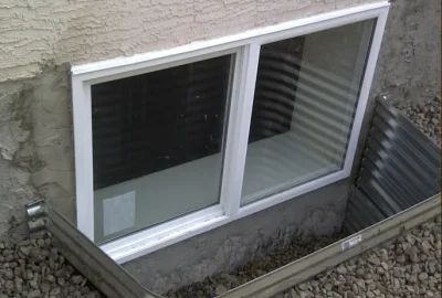 A white-framed basement window set in a stucco wall within a gravel-filled window well.