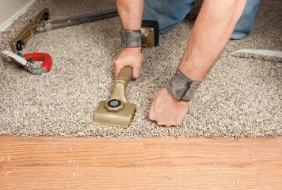 A person uses a carpet kicker tool to install beige carpeting onto a wood floor.