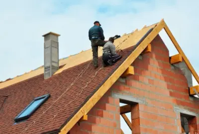 Two construction workers installing red shingles on the roof of a brick house under construction.