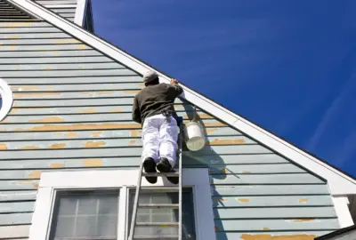 A painter on a ladder scraping peeling blue paint from the siding of a house.